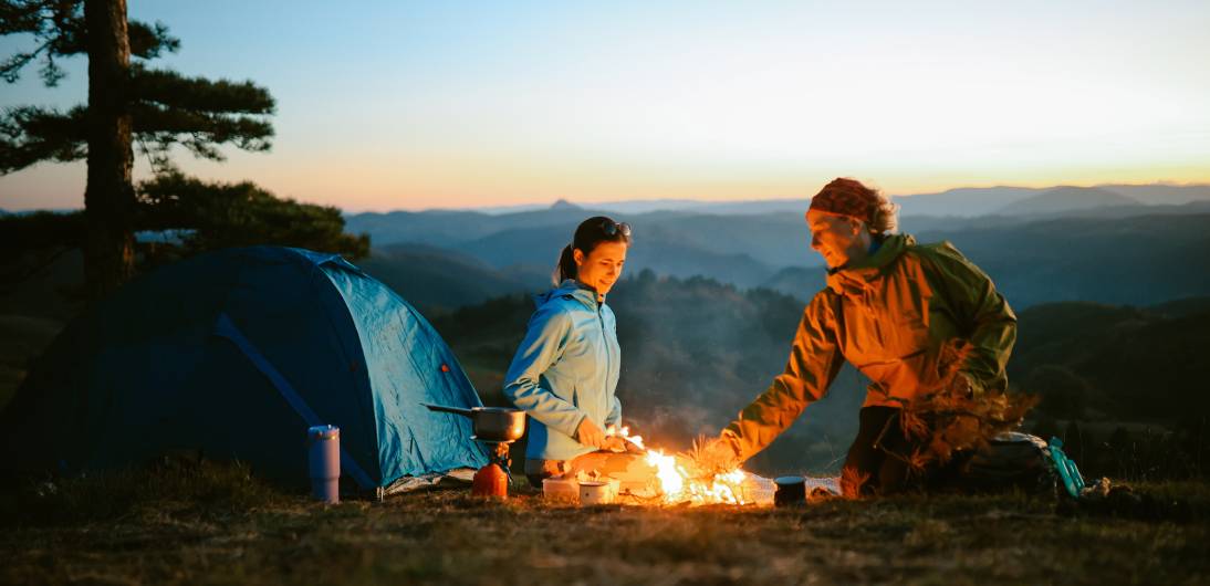 Two people enjoying a campfire beside a tent during sunset on a mountain.