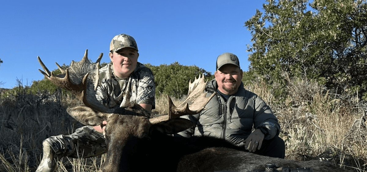 Two hunters posing with a large moose under a clear blue sky.